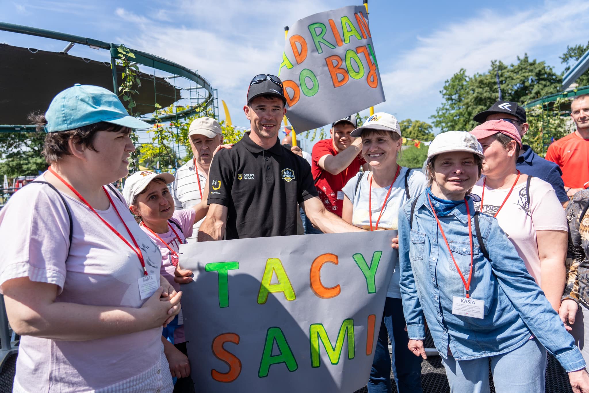 Zdjęcie przedstawia podopiecznych Świetlicy Terapeutycznej Tacy Sami z Adrianem Kosterą podczas startu zawodnika w wyzwaniu 365 Triathlon. Kilka uśmiechniętych osób trzyma dużą kartkę z napisem 'Tacy Sami'.