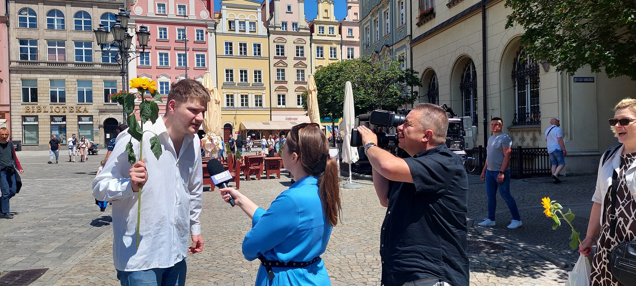 Zdjęcie przedstawia kilka osób na wrocławskim rynku, w tym podopieczny trzymający słonecznik i przemawiający do mikrofonu, który trzyma pani z TVP 3. Obok nich znajduje się kamerzysta, który nagrywa wydarzenie.