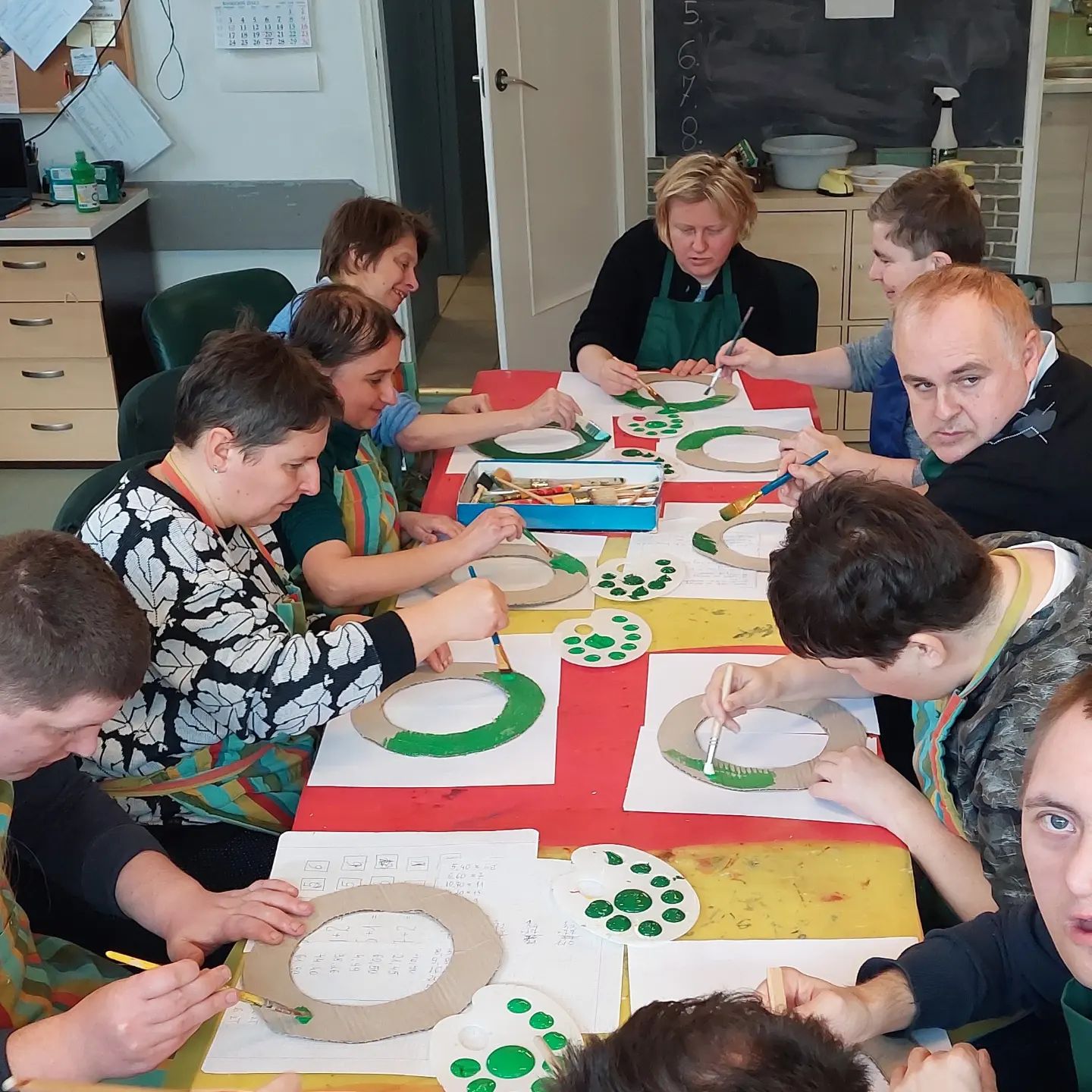 Zdjęcie przedstawia podopiecznych Świetlicy Terapeutycznej Tacy Sami, siedzą przy jednym stole i koncentrują się na malowaniu farbami. Mają na sobie fartuchy, aby nie pobrudzić się.